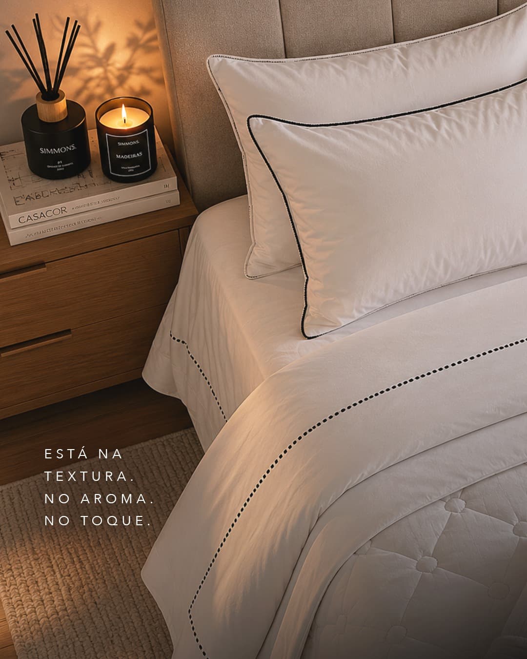 Cama arrumada com jogo de cama branco de vivo preto, à luz de vela, com a legenda “Está na textura. No aroma. No toque.”