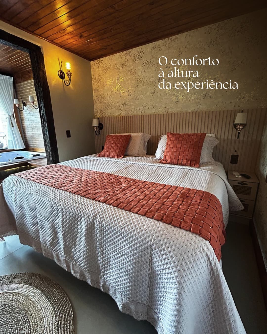 Quarto de pousada com cama de casal arrumada e manta terracota, com a legenda “O conforto à altura da experiência”